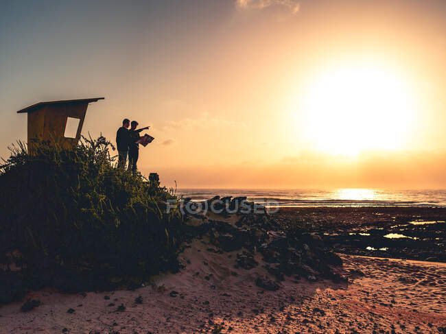 Hombres examinando mapa y apuntando lejos mientras están de pie cerca de refugio en la costa del mar contra el cielo al atardecer en la isla de Fuerteventura, España - foto de stock
