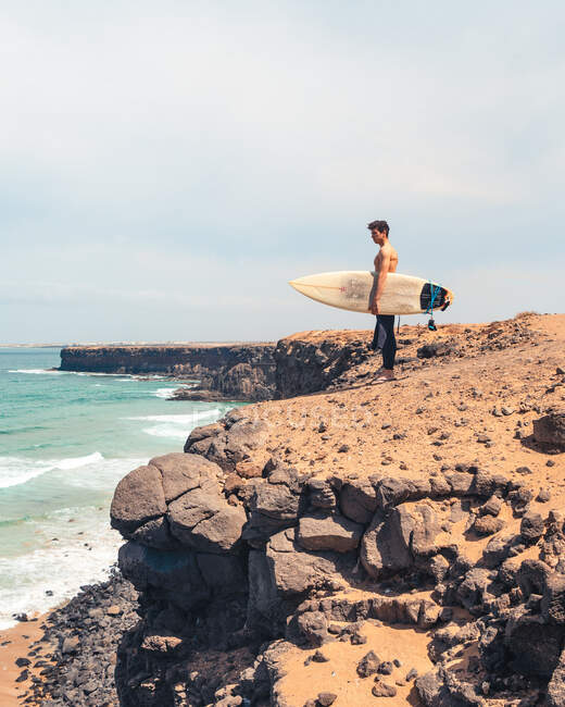 Vista lateral del hombre sin camisa con tabla de surf de pie en la costa pedregosa y admirando el mar ondulante en la isla de Fuerteventura, España - foto de stock