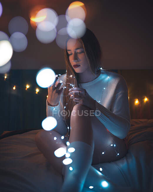 Elegante jovem fêmea em suéter e meias sentado na cama e examinando luzes de fadas brilhantes em acolhedor quarto escuro em casa — Fotografia de Stock