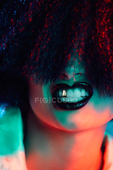 Modelo femenino de corte en peluca sintética y con lápiz labial negro que muestra los dientes bajo luz colorida - foto de stock