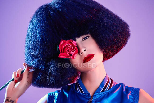 Crop female model in synthetic wig and with red lipstick in a purple background holding a rose — Stock Photo