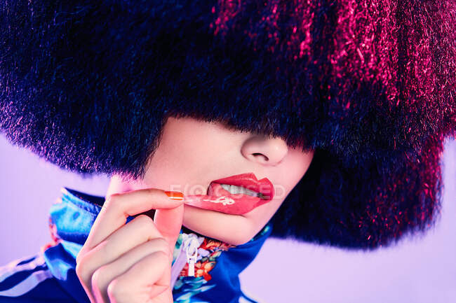 Trendy female in lavish wig removing glossy sticker from lip under magenta light against violet background — Stock Photo
