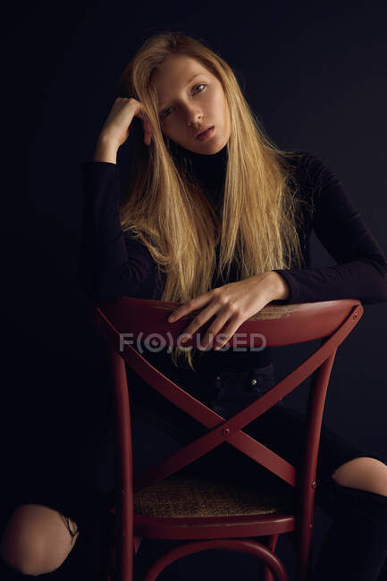Jovem loira em roupas escuras sentada na cadeira contra fundo preto e olhando para longe — Fotografia de Stock