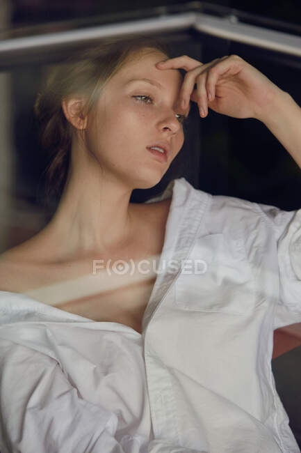 Atractiva modelo femenina joven en camisa blanca con hombro desnudo tocando la cara y mirando hacia otro lado mientras está de pie detrás del vidrio - foto de stock