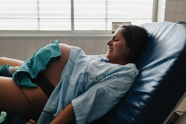Manija de agarre femenina tensa y gruñido de dolor mientras da a luz al bebé en la silla médica en el hospital moderno - foto de stock