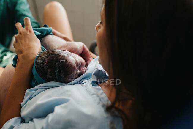 Високий кут веселої дорослої жінки, яка обіймає новонароджену дитину, вкриту кров'ю після народження в кімнаті доставки сучасної лікарні — стокове фото