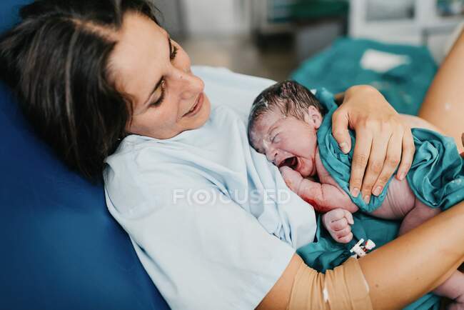 Высокий угол обнимания веселой взрослой женщиной новорожденного ребенка, покрытого кровью, после родов в родильной палате современной больницы — стоковое фото