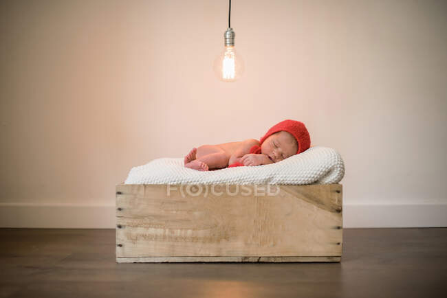 Adorable infant in red hat lying on soft blanket and sleeping in wooden box under glowing light bulb — Stock Photo