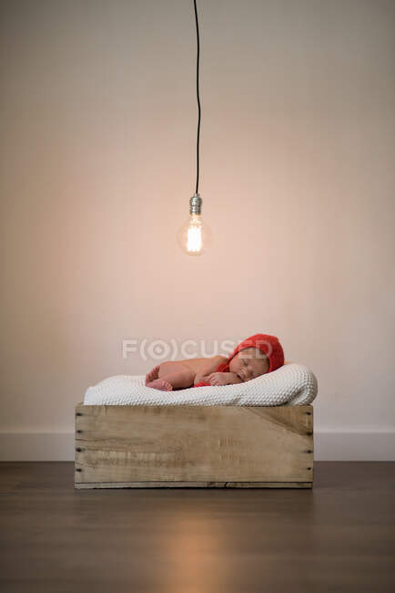 Bebê adorável em chapéu vermelho deitado em cobertor macio e dormindo em caixa de madeira sob lâmpada brilhante — Fotografia de Stock