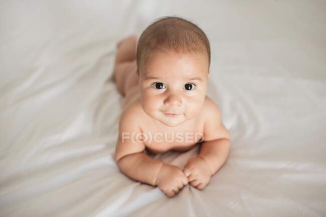 D'en haut adorable bébé souriant et regardant la caméra tout en étant couché sur un lit doux à la maison — Photo de stock