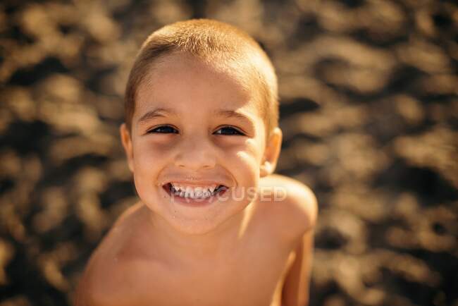 Счастливый мальчик без рубашки улыбается и смотрит в камеру, стоя на песчаном пляже во время заката — стоковое фото