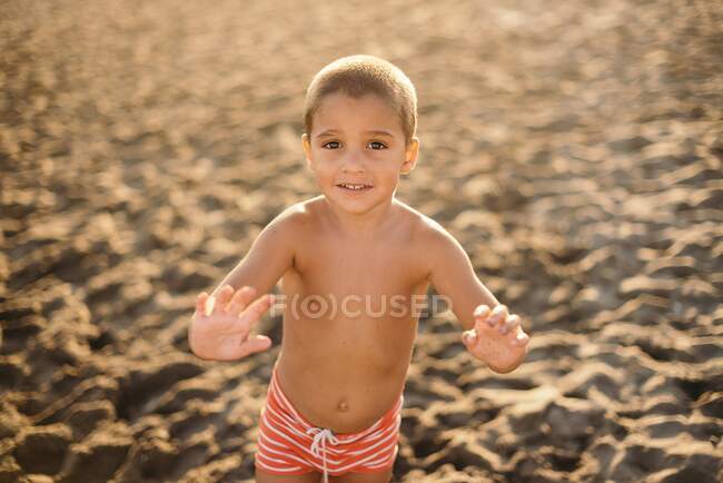 Feliz chico sin camisa sonriendo y mirando a la cámara mientras está de pie en la playa de arena durante la puesta del sol - foto de stock