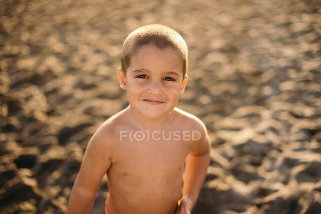 Happy shirtless boy smiling and looking at camera while standing on sandy beach during sunset — Stock Photo