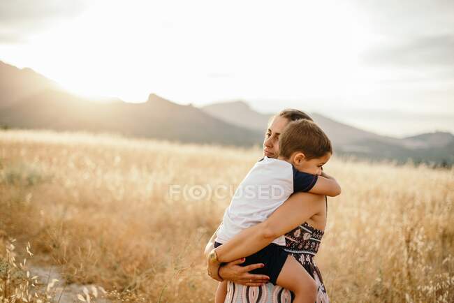 Erwachsene Frau umarmt und trägt Jungen, während sie auf der Wiese vor den Bergen ruht und in der Natur den Sonnenuntergang genießt — Stockfoto