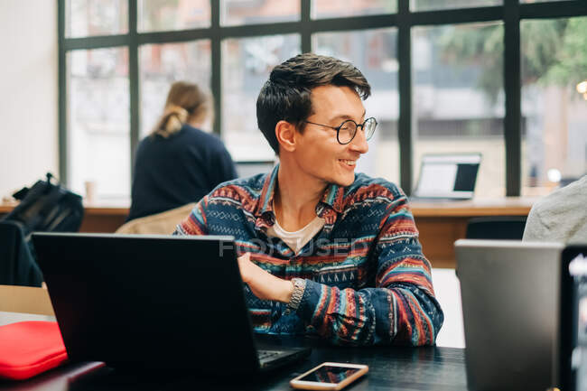 Молодий концентрований чоловік у повсякденному вбранні та окулярах сидить за столом і друкує на ноутбуці під час роботи над проектом у творчій робочій області — стокове фото