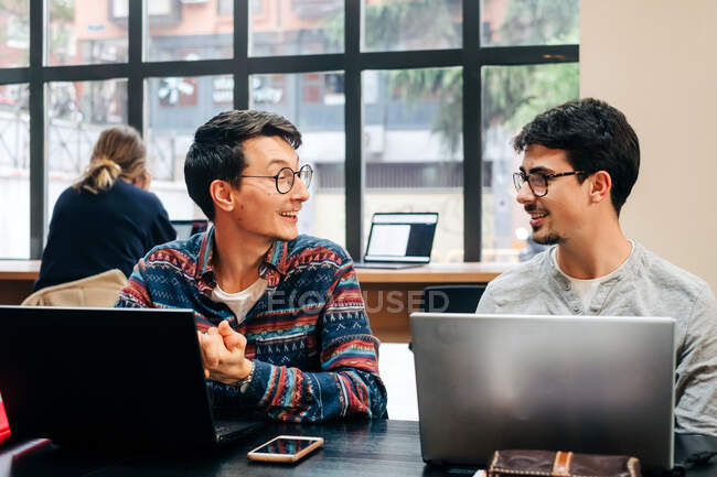 Positive coworkers in casual clothes and eyeglasses sitting at desk and working on project on laptops during workday in modern office — Stock Photo