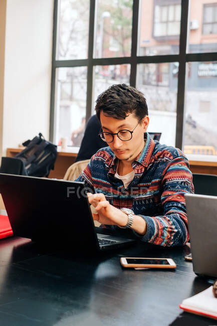 Giovane maschio concentrato in abiti casual e occhiali seduto alla scrivania e digitando sul computer portatile mentre si lavora sul progetto in uno spazio di lavoro creativo — Foto stock
