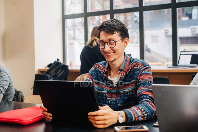 Joven macho concentrado en atuendo casual y anteojos sentados en el escritorio y escribiendo en el portátil mientras trabaja en el proyecto en un espacio de trabajo creativo - foto de stock
