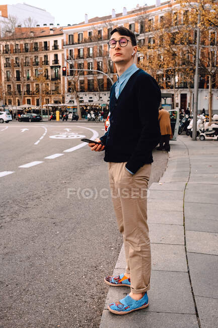 Повне тіло молодого впевненого модного чоловіка в модному вбранні та окулярах зі смартфоном у руці, що стоїть на вулиці — стокове фото