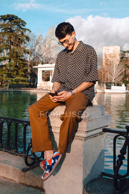 Full length of Asian cheerful trendy guy in stylish wear and eyeglasses surfing smartphone and looking at screen while sitting on bridge railing and listening to music on air pods — Stock Photo
