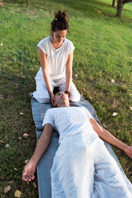 From above of calm couple meditating while man lying on mat with eyes closed and woman sitting on knees and holding partner by head during looking down in green meadow — Stock Photo