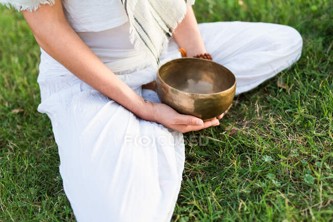 Vista lateral de calma jovem do sexo masculino em branco desgaste sentado sobre os joelhos e segurando tibetano cantando tigela em mãos enquanto vai ioga e relaxante no gramado no dia de verão — Fotografia de Stock