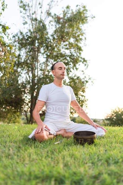 Vista lateral de la calma masculina joven en ropa blanca sentado en las rodillas y sosteniendo el cuenco de canto tibetano en las manos mientras va a yoga y se relaja en el césped en el día de verano - foto de stock