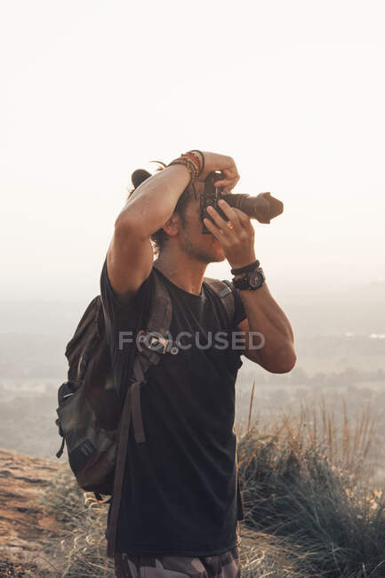 Side view of young male traveler in casual clothes with haversack standing on edge of cliff and taking picture of majestic landscape of green forest during sunset — Stock Photo