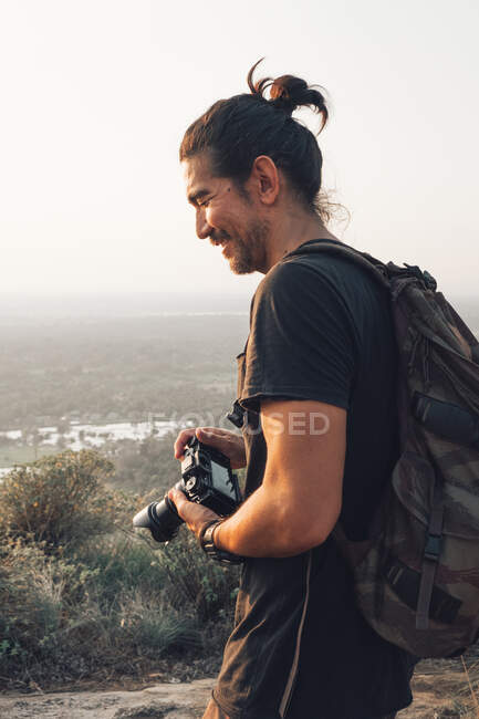 Side view of young cheerful male traveler in casual clothes with haversack standing on edge of cliff and taking picture of majestic landscape of green forest during sunset — Stock Photo