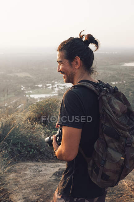 Side view of young cheerful male traveler in casual clothes with haversack standing on edge of cliff and taking picture of majestic landscape of green forest during sunset — Stock Photo