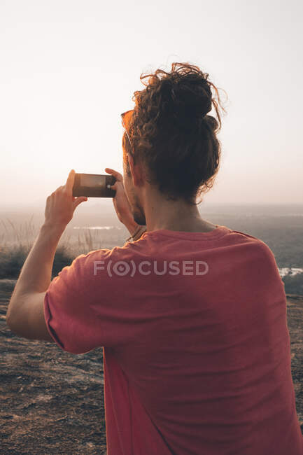Вид ззаду молодого невпізнаваного чоловіка-мандрівника в повсякденному одязі, що стоїть на краю скелі і фотографує величний пейзаж зеленого лісу на безхмарне блакитне небо під час заходу сонця — стокове фото