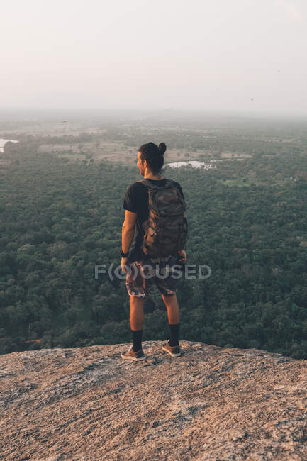 Back view of young unrecognizable male traveler in casual clothes with haversack standing on edge of cliff against majestic landscape of green forest and cloudless blue sky during sunset — Stock Photo