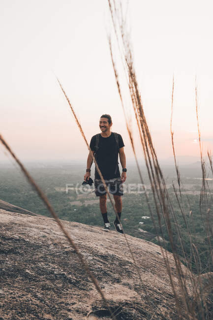 Вид спереди на молодого путешественника в повседневной одежде с повозкой, стоящей на краю скалы против величественного ландшафта зеленого леса и безоблачного голубого неба на закате — стоковое фото
