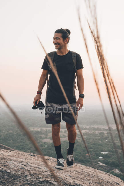 Молодой веселый мужчина-путешественник в повседневной одежде с фотокамерой, стоящей на краю скалы и фотографирующей величественный пейзаж зеленого леса на закате — стоковое фото