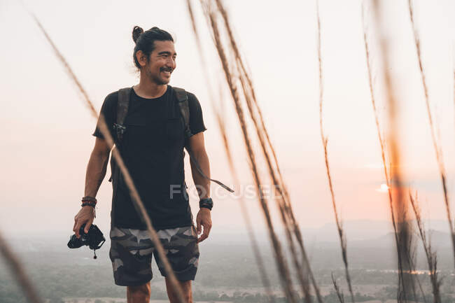 Jovem viajante masculino alegre em roupas casuais com câmera fotográfica em pé na borda do penhasco e tirar fotos da majestosa paisagem da floresta verde durante o pôr do sol — Fotografia de Stock