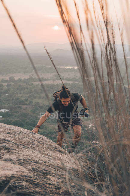 Анонімний чоловічий турист у повсякденному одязі та рюкзаку, що піднімається скелястою скелею та тримає фотоапарат в руці, дивлячись вниз на тлі заходу сонця — стокове фото