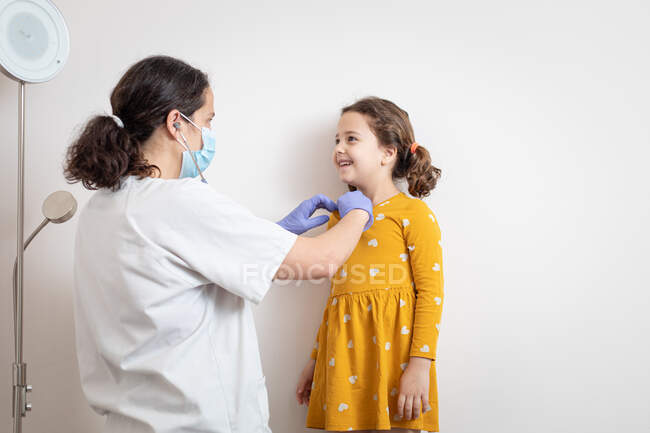 Side view of female doctor in white uniform and in latex gloves and protective mask examining with stethoscope lungs of small girl — Stock Photo