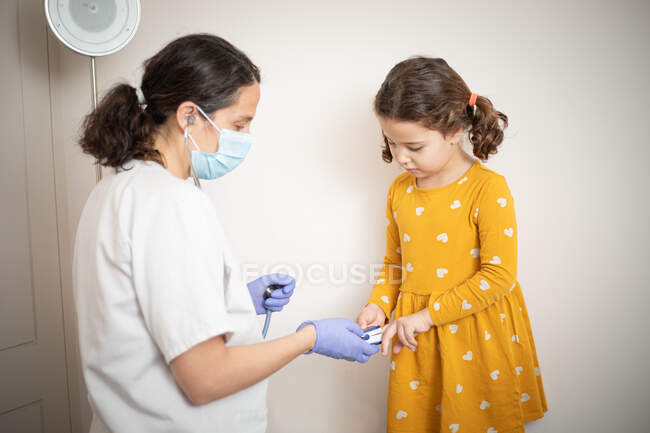 Un médico con guantes de látex usando un puño para examinar la presión arterial de una niña con un vestido amarillo casual - foto de stock