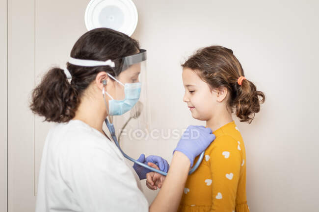 Vista lateral de la doctora en uniforme blanco y en guantes de látex y máscara protectora examinando con estetoscopio los pulmones de una niña pequeña - foto de stock