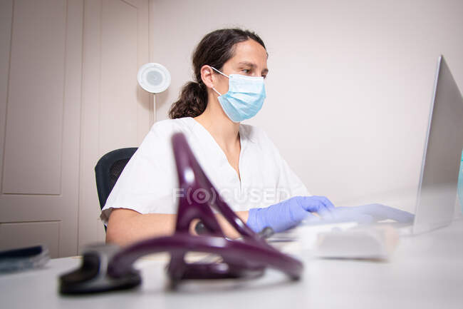 Серйозна молода жінка-лікар в білій формі та медична маска, що працює на ноутбуці в латексі, сидить за столом у сучасній клініці — стокове фото