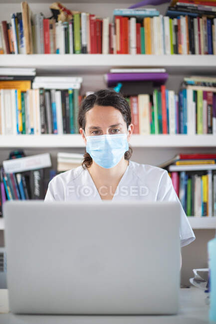 Jeune médecin sérieuse portant un uniforme blanc et un masque médical travaillant sur un ordinateur portable en gants de latex assis au bureau dans une clinique moderne — Photo de stock