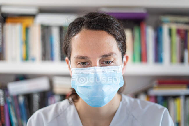 Médica jovem positivo em uniforme branco e estetoscópio vestindo máscara médica protetora em pé no escritório médico moderno e olhando para a câmera — Fotografia de Stock