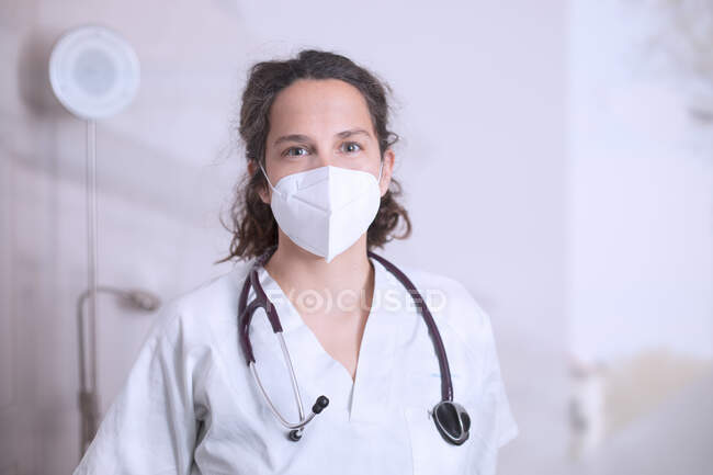 Positive junge Ärztin in weißer Uniform und Stethoskop mit medizinischer Schutzmaske steht in moderner Arztpraxis und blickt in die Kamera — Stockfoto