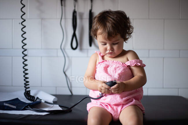 Цікава чарівна маленька дівчинка в повсякденній рожевій сукні сидить на лавці в медичній кімнаті і грає з тонометром під час відвідування лікаря — стокове фото
