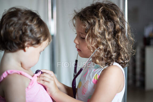 Seitenansicht eines kleinen Mädchens mit lockigem Haar bei der Untersuchung der Lungen eines anderen kleinen Mädchens mit Stethoskop während des Besuchs einer modernen Klinik — Stockfoto