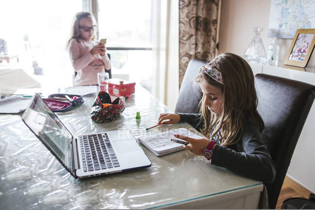 Chica inteligente tomando notas en el cuaderno mientras está sentado en la mesa cerca de la hermana y haciendo la tarea de la computadora portátil en casa - foto de stock