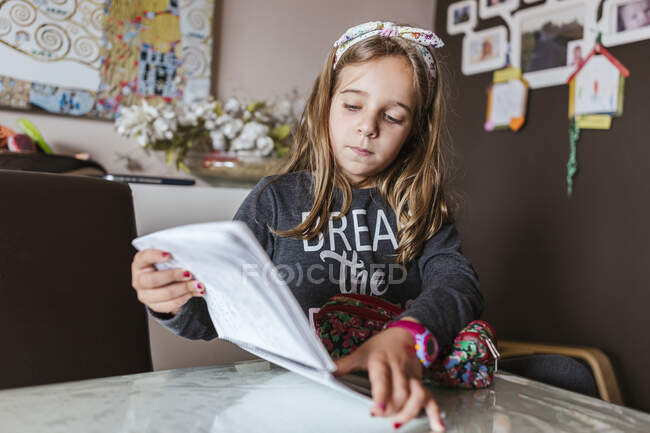 Menina sentada à mesa e ler notas no bloco de notas ao fazer a tarefa de casa em casa — Fotografia de Stock