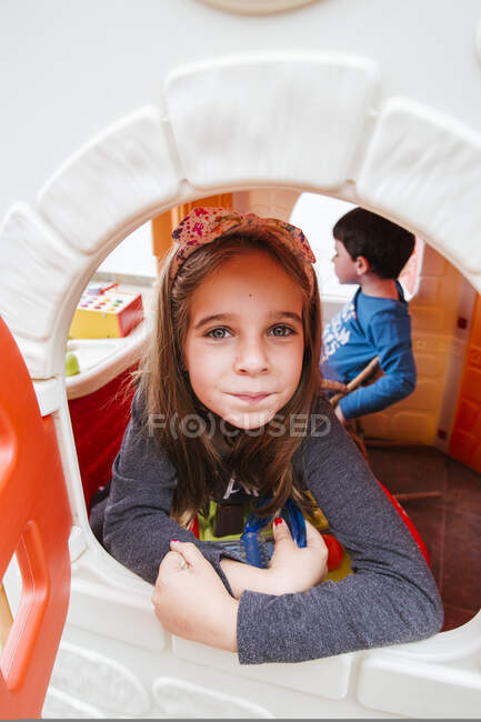 Маленькая девочка смотрит в камеру через окно игрушечного домика, играя с братом дома — стоковое фото