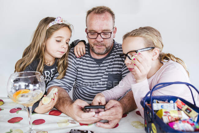 Meninas comendo banana e navegando nas mídias sociais no smartphone enquanto passam tempo com o pai na mesa em casa — Fotografia de Stock