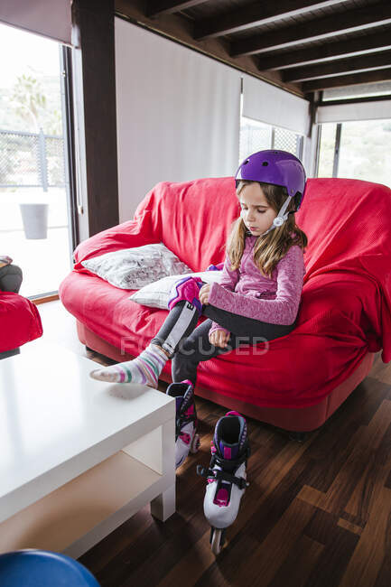 Mädchen mit buntem Helm sitzt zu Hause auf Couch und setzt sich Knieschützer auf, bevor sie Rollschuh fährt — Stockfoto